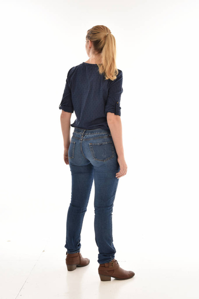 po celé délce portrét blond dívka, která nosí jednoduché modrou košili a džínách, stojící pozice směrem od kamery. izolované na bílém pozadí. - Fotografie, Obrázek