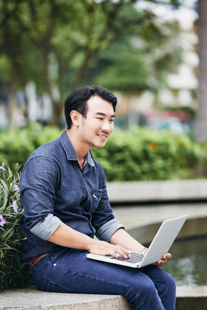 Veselá vietnamské mladý muž sedí venku a čtení e-mailů na laptop - Fotografie, Obrázek