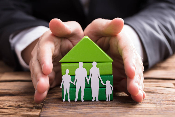 Businessperson's Hand Protecting House Model And Family Paper Cut Out On Wooden Desk - Foto, Imagen