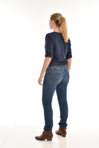 カメラから離れて直面しているポーズに立っているシンプルなブルーのシャツとジーンズを身に着けている金髪の女の子の完全な長さの肖像画。白い背景に分離. - 写真・画像