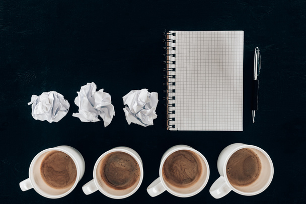 vista superior do bloco de notas em branco com papéis amassados e xícaras de café em fileira isolado em preto
 - Foto, Imagem