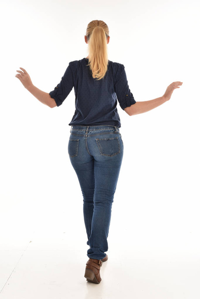 full length portrait of blonde girl wearing simple blue shirt and jeans, standing pose facing away from the camera. isolated on white background. - Photo, Image