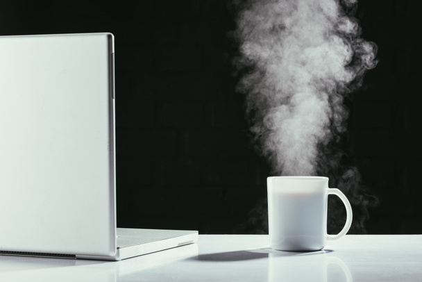 portatile con tazza di tè fumante sul tavolo isolato su nero
 - Foto, immagini