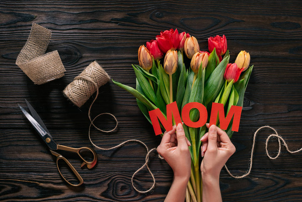 partial view of woman with mom lettering in hands and bouquet of tulips on wooden tabletop, mothers day concept - Foto, Bild