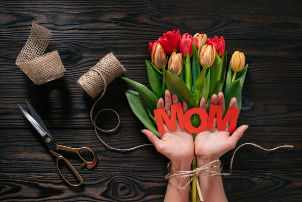 partial view of woman with mom lettering in hands and bouquet of tulips on wooden tabletop, mothers day concept - Фото, зображення