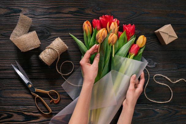 partial view of woman wrapping bouquet of tulips into wrapping paper on wooden tabletop - Foto, afbeelding