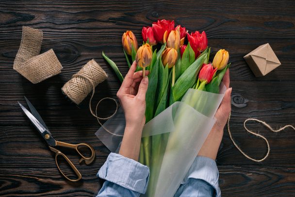 partial view of woman wrapping bouquet of tulips into wrapping paper on wooden tabletop - Photo, Image