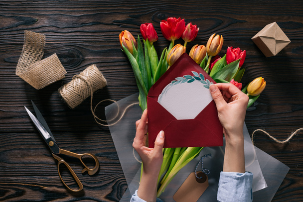 Schnappschuss einer Frau mit Postkarte in den Händen und Tulpen in Packpapier auf Holztischplatte - Foto, Bild