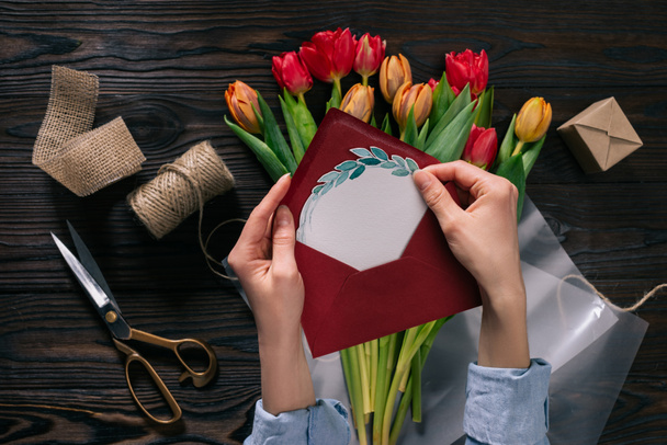 cropped shot of woman with postcard in hands and tulips in wrapping paper on wooden tabletop - Photo, Image
