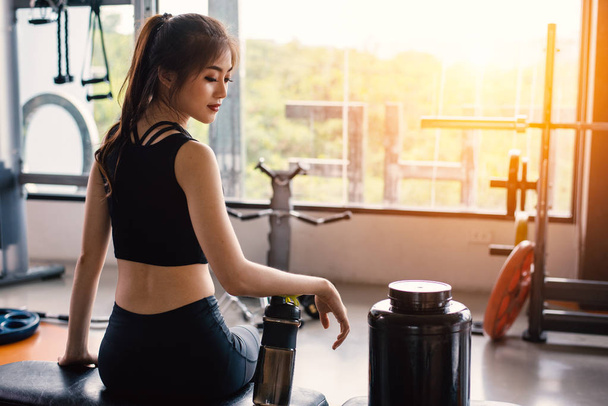 Sport woman sitting and resting after workout or exercise in fitness gym with protein shake or drinking water on floor. Relax concept. Strength training and Body build up theme. Warm and cool tone - Photo, image
