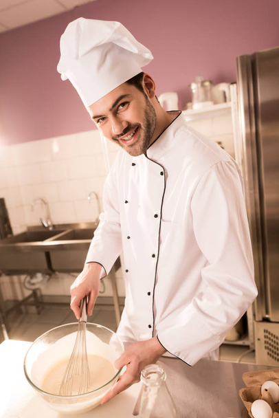 πορτρέτο του χαμογελώντας ζαχαροπλάστη στο καπέλο του σεφ κάνοντας ζύμη σε κουζίνα εστιατορίου - Φωτογραφία, εικόνα