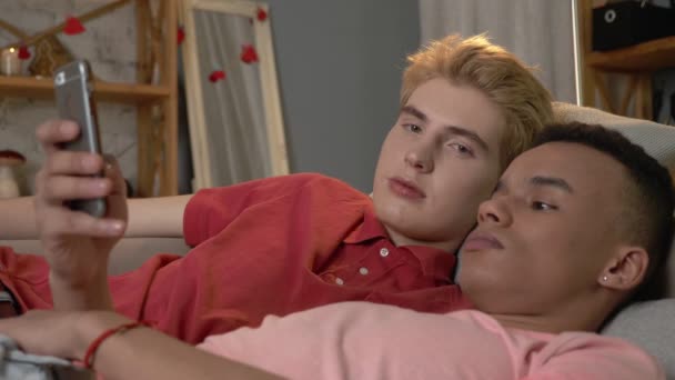 Un par de jóvenes homosexuales multinacionales yacían en el sofá y usaban un smartphone. Homeliness, gay, love, young LGBT family concept 60 fps
 - Imágenes, Vídeo