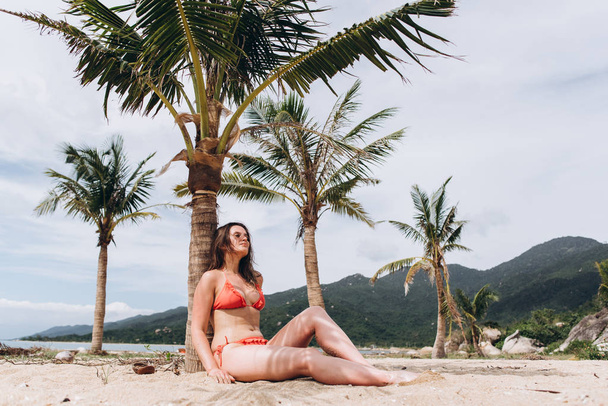View of woman wearing red bikini sitting on sand near palms and posing on mountains background. - Photo, Image
