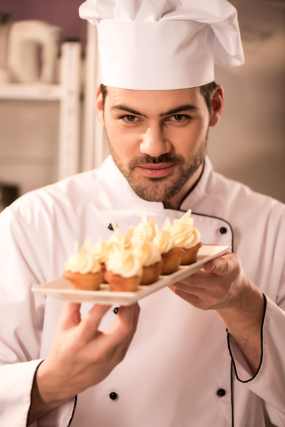 retrato de joven pastelero mirando cupcakes en plato en las manos
 - Foto, Imagen