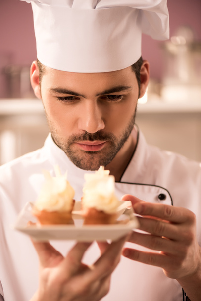 portrait of focused confectioner looking at cupcakes on plate in hands - Foto, immagini