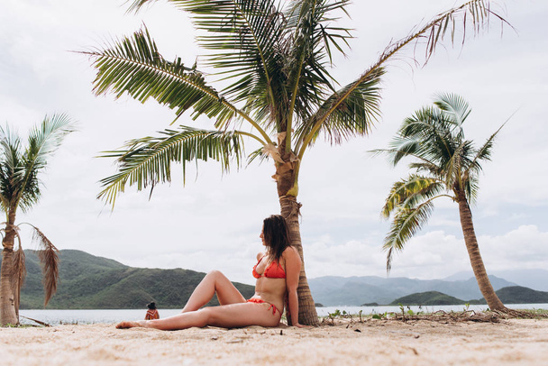 View of woman wearing red bikini sitting on sand near palms and posing on mountains background. - Foto, Bild