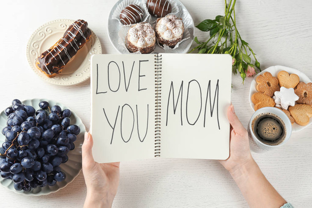 Woman holding notebook with words "Love you mom" at table. Mother's day breakfast - Foto, Imagen