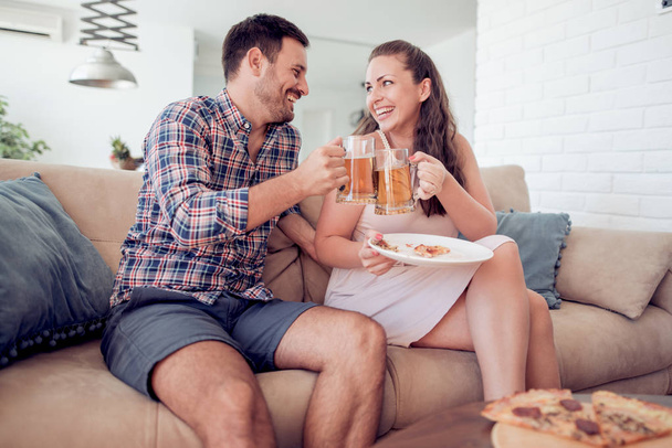 ビールを飲み、ピザを食べて、テレビを見ている美しい若いカップル。屋内で. - 写真・画像