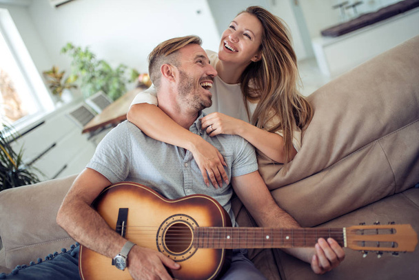 Milenci v obývacím pokoji. Mladý pár v lásce sedí na pohovce, zatímco mladík hrál na kytaru v obývacím pokoji. - Fotografie, Obrázek