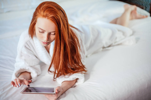 beautiful woman wearing bathrobe and using digital tablet while relaxing on bed - Photo, image