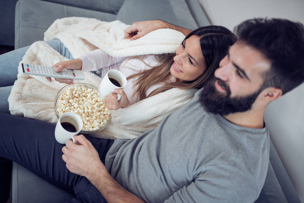 Jong koppel ontspannen in hun appartement, liggend op de bank kijken naar een film en eten popcorn. - Foto, afbeelding