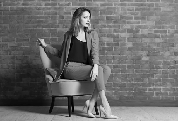 Beautiful young stylish woman sitting in armchair near brick wall - 写真・画像