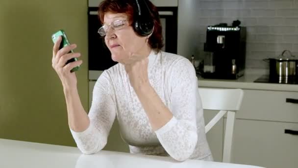 Starsza kobieta słucha muzyki na słuchawkach z telefonu, gesticulates energicznie i tańczy - Materiał filmowy, wideo