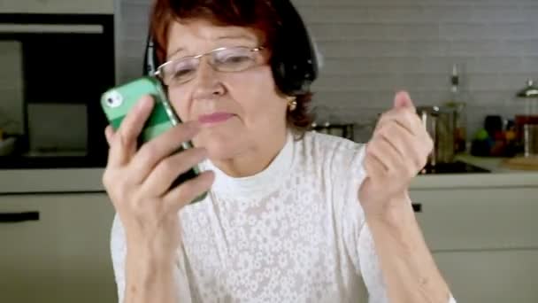 An elderly woman listens to music on headphones from the phone, gesticulates energetically and dances - Footage, Video