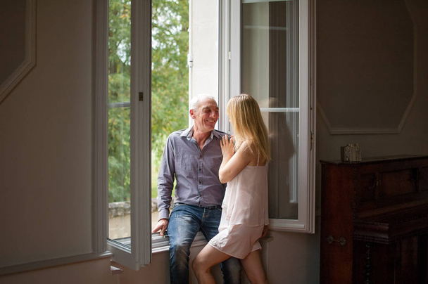 Hombre mayor en pantalones vaqueros y camisa abrazando a su joven esposa rubia de pie cerca de la ventana en su casa durante el verano
. - Foto, imagen
