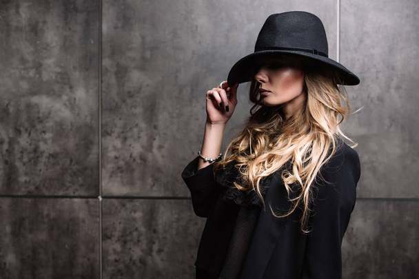 Beautiful young mysterious blond girl in black hat and black jacket on gray background. Eyes are covered with a hat. Manicure - long red nails, nail polish. Fashion, beauty. - 写真・画像