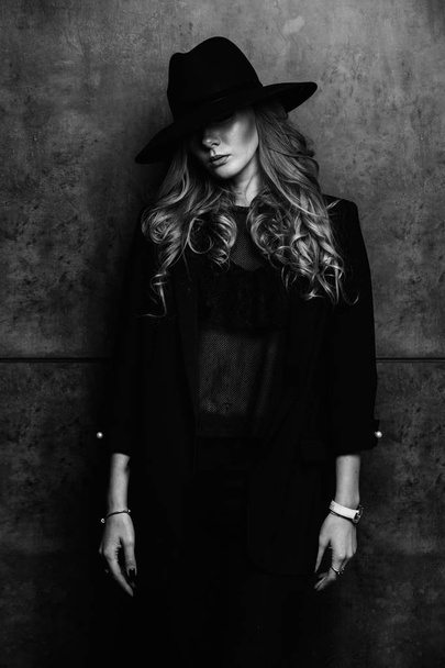 Beautiful young mysterious blond girl in black hat and black jacket on gray background. Eyes are covered with a hat. Manicure - long red nails, nail polish. Fashion, beauty. - Fotó, kép