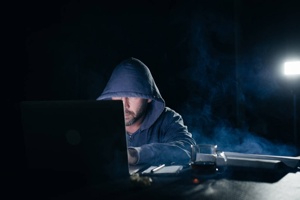 een mannelijke hacker zit in een donkere kamer en iets typt op een laptop - Foto, afbeelding