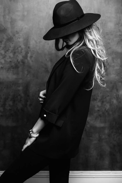 Beautiful young mysterious blond girl in black hat and black jacket on gray background. Eyes are covered with a hat. Manicure - long red nails, nail polish. Fashion, beauty. - 写真・画像