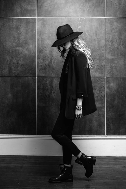 Beautiful young mysterious blond girl in black hat and black jacket on gray background. Eyes are covered with a hat. Manicure - long red nails, nail polish. Fashion, beauty. - Φωτογραφία, εικόνα