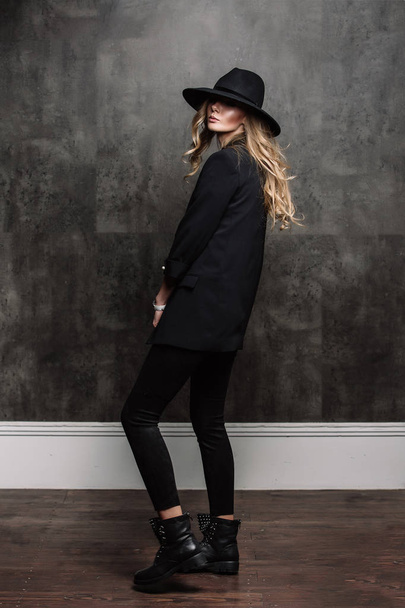 Beautiful young mysterious blond girl in black hat and black jacket on gray background. Eyes are covered with a hat. Manicure - long red nails, nail polish. Fashion, beauty. - Zdjęcie, obraz