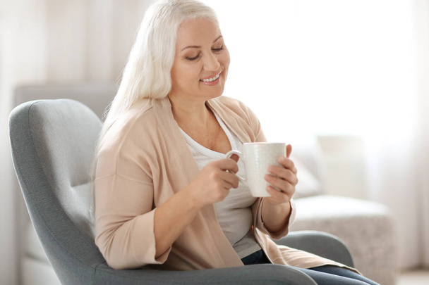 Happy mature woman drinking tea while sitting in armchair at home - Foto, Bild