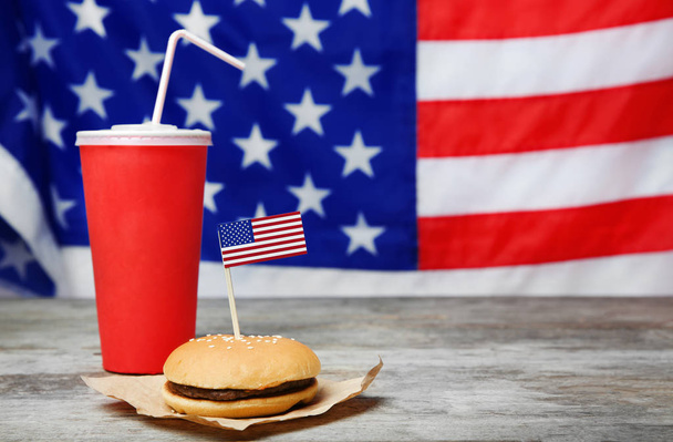 Tasty hamburger and cup with drink on wooden table against blurred American flag - Zdjęcie, obraz