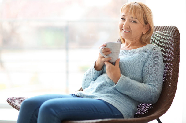Happy mature woman drinking tea while sitting in armchair at home - Foto, Imagen