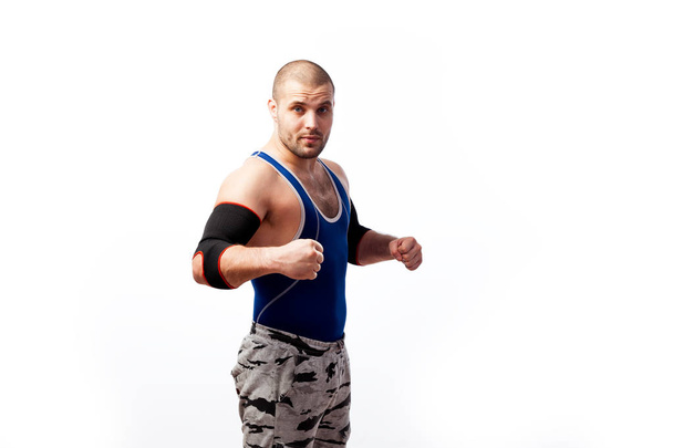 Sportieve jongeman worstelaar in een blauwe tank top, sport broek en elleboog pads staat en heeft zijn handen in vuisten op een witte achtergrond van de geïsoleerde, zijaanzicht - Foto, afbeelding
