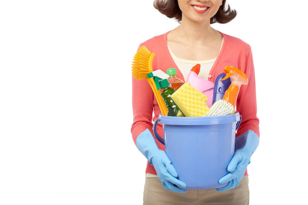 smiling housewife holding bucket with cleaning utensils in hands while standing against white background - Photo, Image