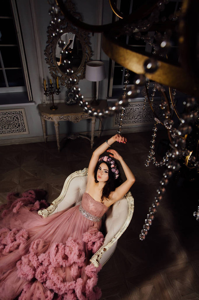 A beautiful slender brunette woman in a pink dress with flounces is sitting on a blue velvet armchair - Fotografie, Obrázek