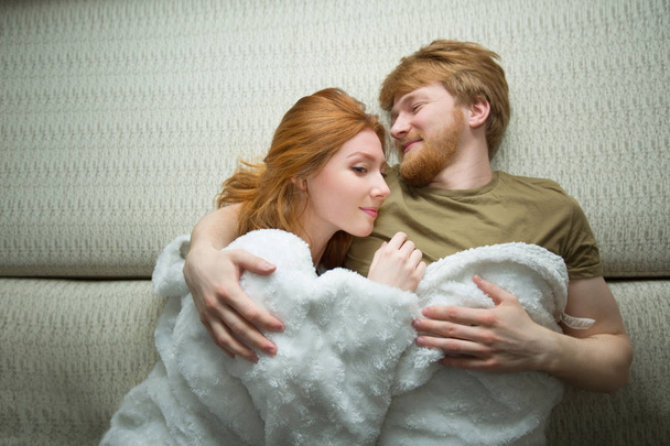 happy young couple guy with a girl with red hair lying on a sofa under a blanket - Фото, зображення