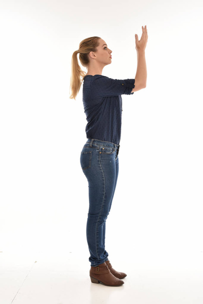 Ganzkörperporträt eines Mädchens, das einfaches blaues Hemd und Jeans trägt und im Profil vor weißem Studiohintergrund steht. - Foto, Bild