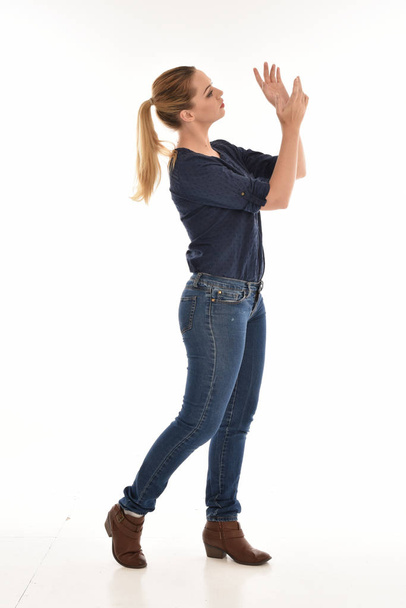 full length portrait of girl wearing simple blue shirt and jeans, standing in profile against a white studio background. - 写真・画像