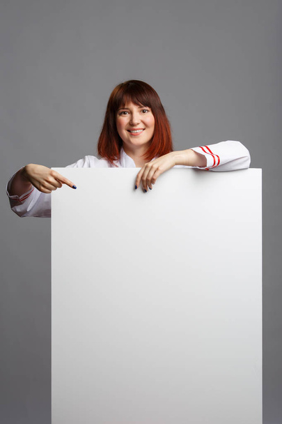 Photo of smiling female cook in white robe pointing finger at empty sheet for inscription - Valokuva, kuva