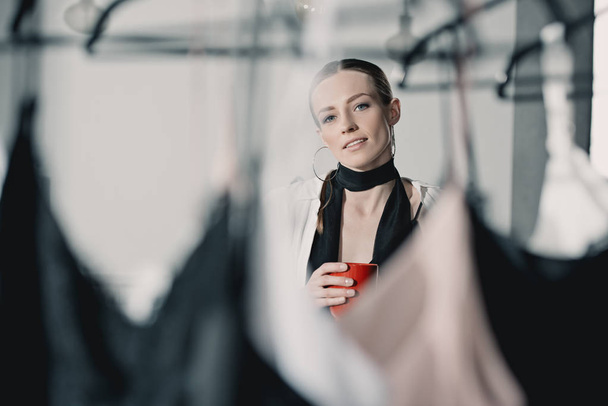 stylish young woman with coffee mug looking at clothes on rack - Fotó, kép