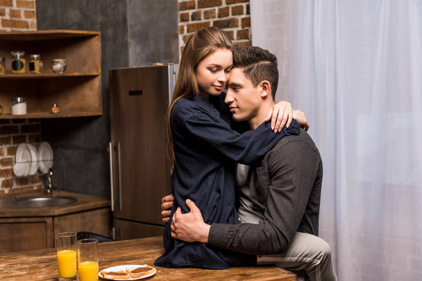 girlfriend sitting on table in kitchen and hugging boyfriend - Foto, imagen