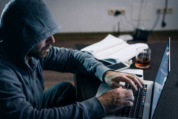 a male hacker is sitting in a dark room at the table and something is typing on a laptop - Foto, Imagem