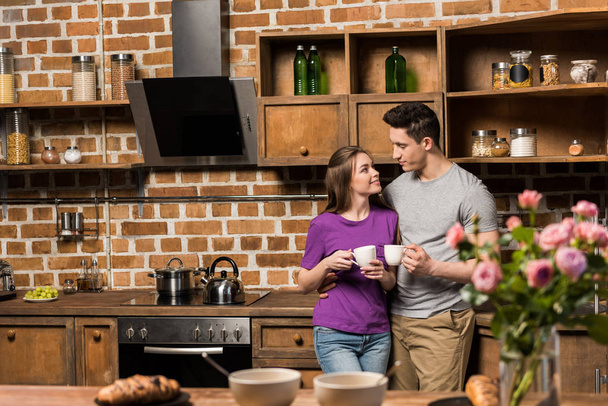 couple looking at each other in kitchen and holding cups of coffee - Photo, Image