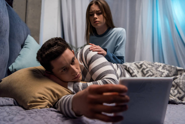 přítel sleduje něco na tabletu a přítelkyně sedí rozzlobený na posteli - Fotografie, Obrázek